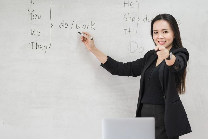 Kadın öğretmen, beyaz tahtada "do/work" yazarken öğrencilere işaret ediyor. Gülümseme ve profesyonel giyim, öğretim ortamına katkı sağlıyor.