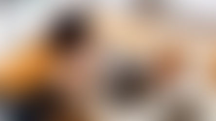 Woman in yellow shirt leans over a kitchen sink filled with dishes, hands on face. Modern kitchen setting, neutral tones, overwhelmed mood.