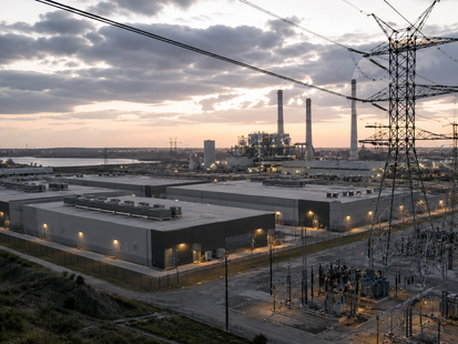 Ein grossflächiges Rechenzentrum mit flachem Dach liegt direkt neben einem Umspannwerk und Hochspannungsleitungen. Im Hintergrund ist ein Kraftwerk zu sehen. Das Bild visualisiert den wachsenden Energiebedarf künstlicher Intelligenz und zeigt, wie Rechenzentren zunehmend zur direkten Schnittstelle zwischen Technologie, Stromversorgung und strategischer Infrastruktur werden.