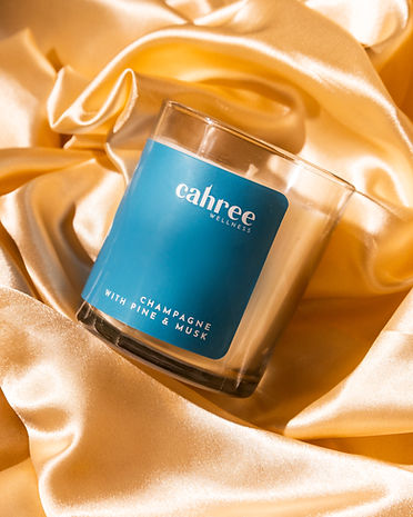 Cahree Champagne with Pine & Musk luxury soy candle in blue glass jar, resting on golden satin fabric for an elegant, warm-toned composition