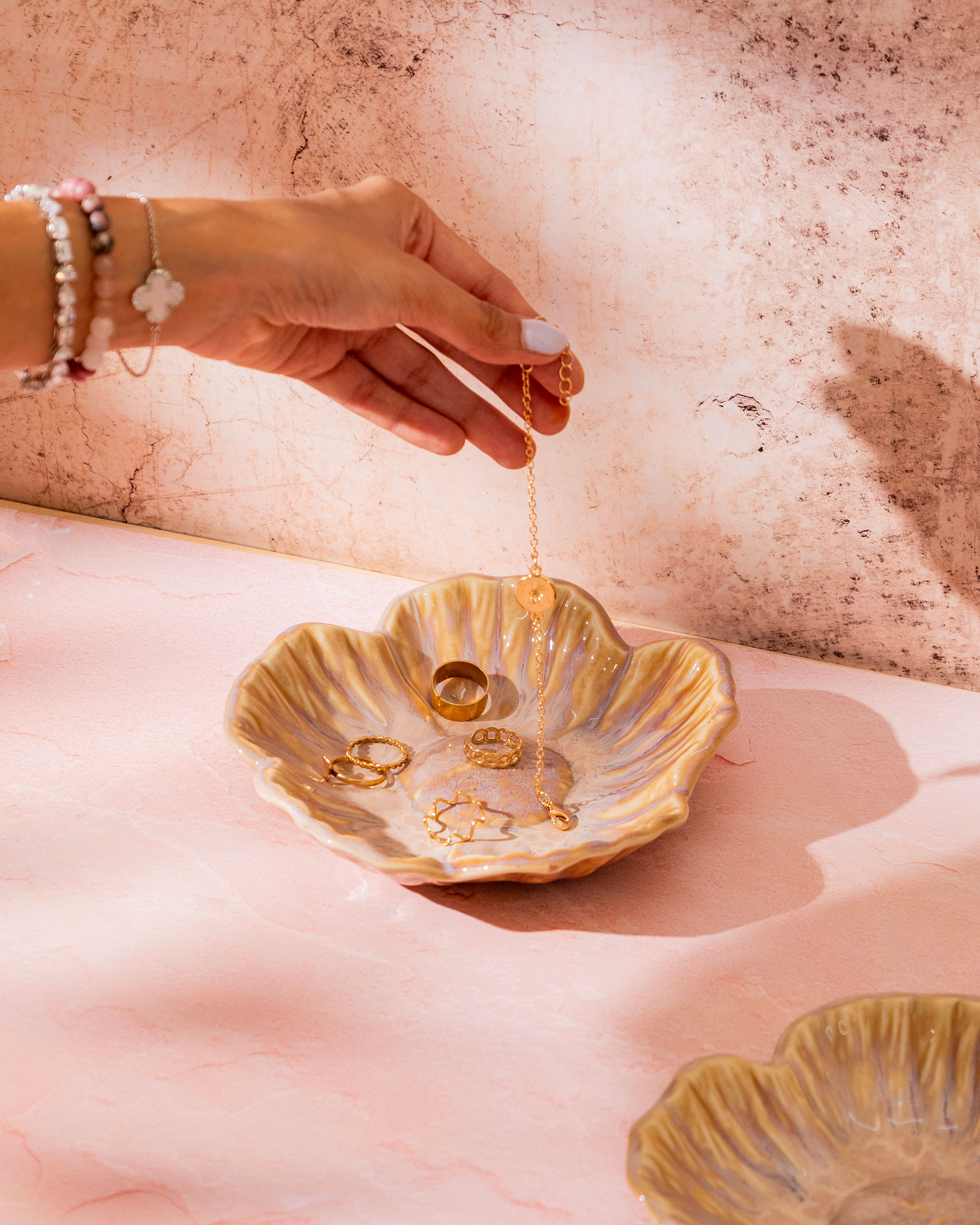 Golden ceramic petal-shaped trinket tray holding rings and jewellery on a soft pink surface in a minimal setting.