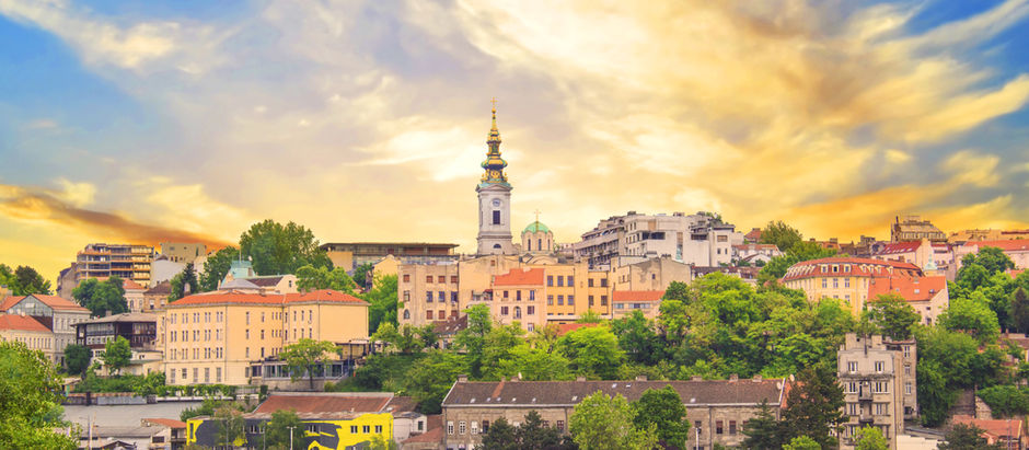 Boats on a river with colorful buildings and a tower in the background. The sky is vibrant with orange and blue hues, creating a serene mood.
