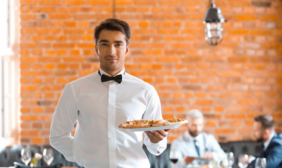 Waiter in a white shirt and bow tie holds a pizza in a restaurant with brick walls. Two men are blurred in the background.