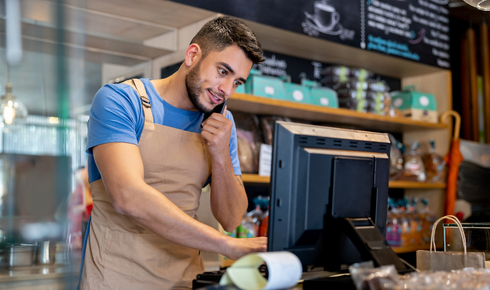 Man in a blue shirt and apron in a bakery, talking on the phone and using a computer. Shelves with packaging and menu board behind.