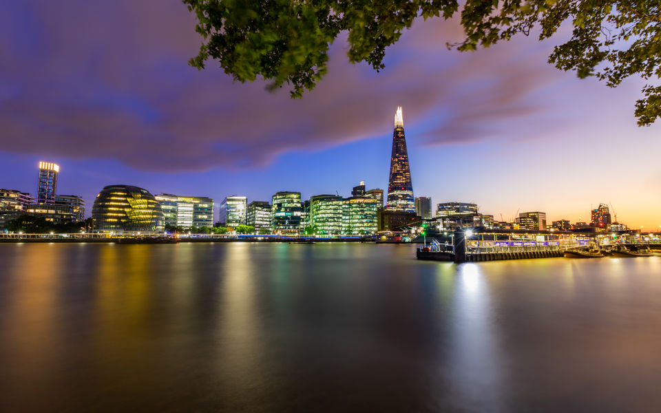 A mesmerizing view of London's skyline at twilight, with the iconic Shard towering above the city. The river Thames glistens with reflections of the illuminated buildings, creating a captivating cityscape.