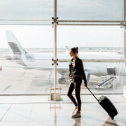 Woman walking in airport with luggage being pulled behind her in front of a glass wall with airplane in the background before a long flight.