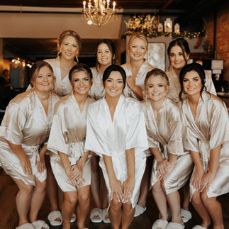 Bride and bridesmaids wearing matching satin robes, smiling together inside a historic brick wedding venue during bridal getting ready moments.