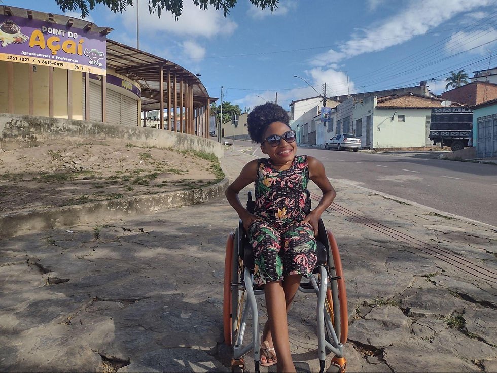 Na imagem há uma mulher negra, usuária de cadeira de rodas, posicionada no centro, ela sorri e apoia os braços na cintura usando um vestido preto com flores rosas. No fundo da paisagem, há uma placa escrito Point do Açaí.