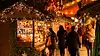 People browse a festive outdoor holiday market stall, reminiscent of Cap Ferret in the off season, decorated with fairy lights, greenery, and baubles at night. Colourful gifts create a warm and cheerful atmosphere despite the tranquil time of year.