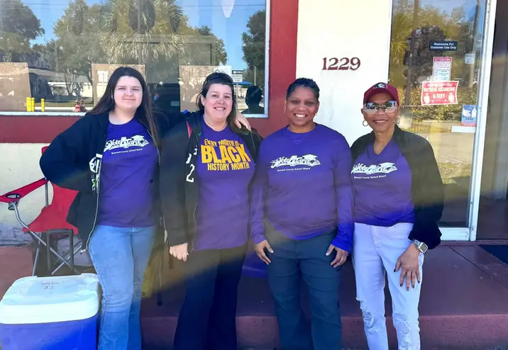 Four women stand together and smile outside a building with the number 1229. Three are wearing matching purple shirts, and one wears a maroon cap and sunglasses. A cooler and a red chair are visible beside them.