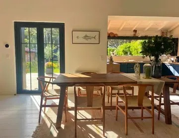 Sunlit dining area with a wooden table and six chairs, a light rug, glass doors leading outside, and a kitchen in the background. A framed fish print hangs on the wall and plants decorate the table and kitchen.