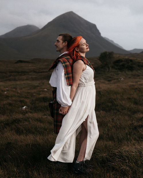 Couple holding hands in the Scottish Highlands with a dramatic mountain behind them, the bride wearing a flowing white dress and tartan shawl.