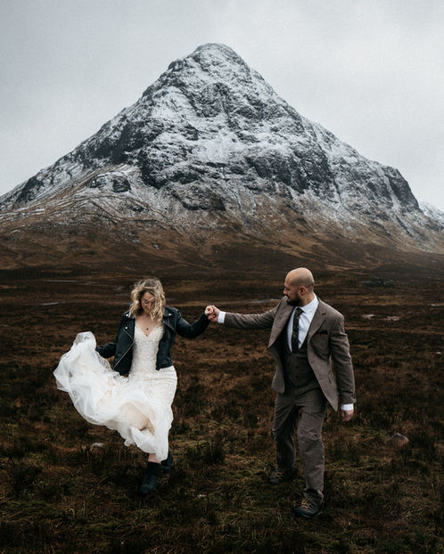 Groom holding bride's hand underneath snow-capped mountain in Glencoe, Scotland