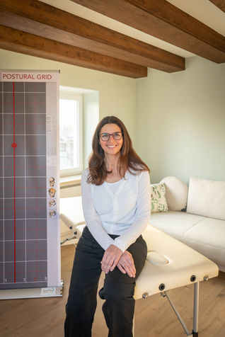 Lachende Frau sitzt auf Behandlungsliege, "POSTURAL LINES" Chart im Therapieraum.