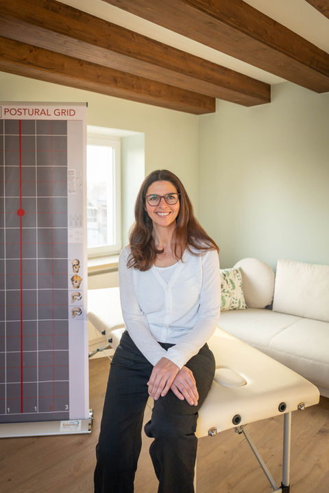 Lachende Frau sitzt auf Behandlungsliege, "POSTURAL LINES" Chart im Therapieraum.