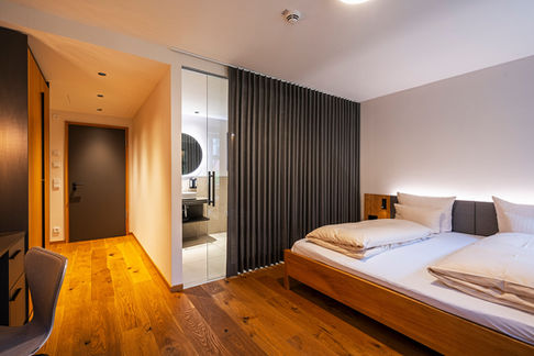 Modernes Hotelzimmer mit Holzboden und Bad hinter Glas. Ein Dunkler Vorhang vor dem Badezimmer. 
