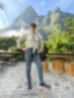 A woman in casual attire stands smiling on a stone patio with lush mountains and blue sky in the background, surrounded by tree stump tables.