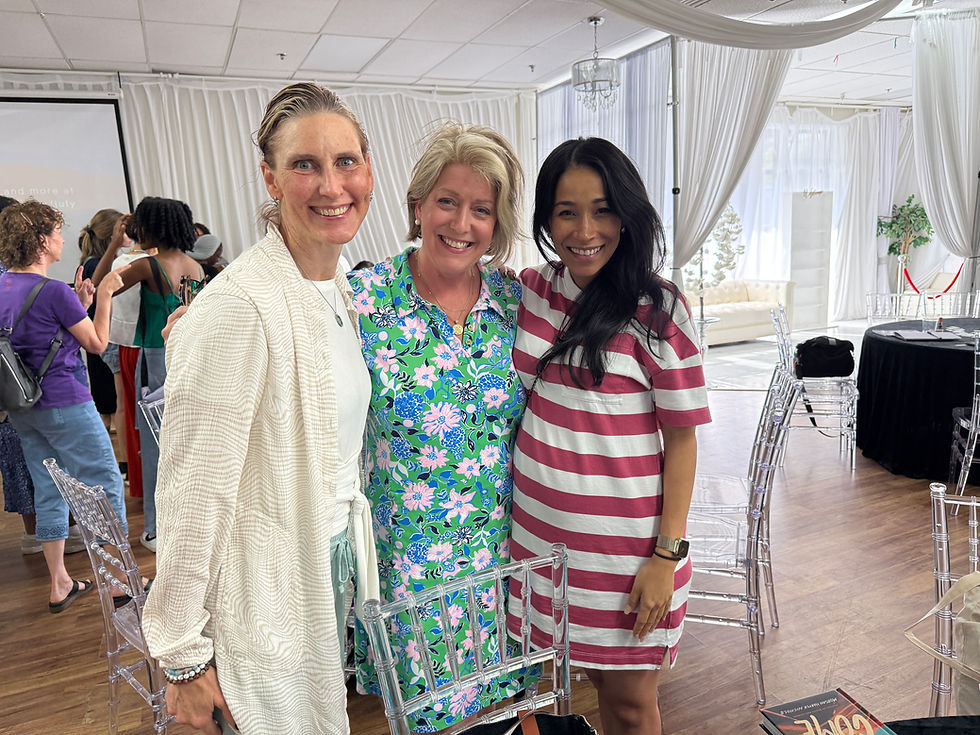 Three women smiling in a decorated room with white drapes. One wears a white cardigan, another a floral dress, and the third pink stripes.