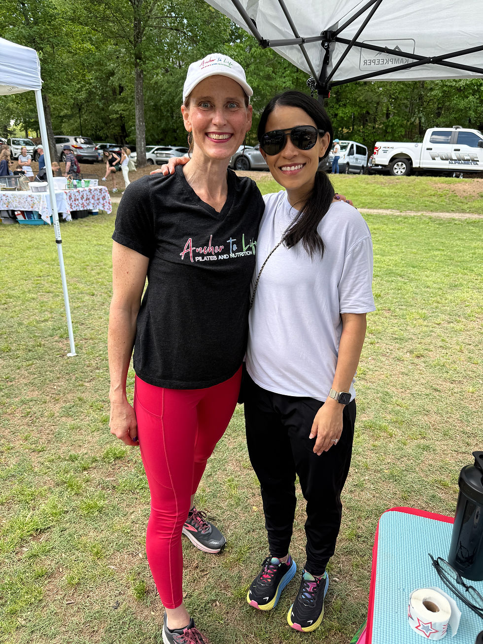 Two women smile outdoors under a canopy. One wears a black tee and red leggings, the other in a white tee. Cars and tables are in the background.
