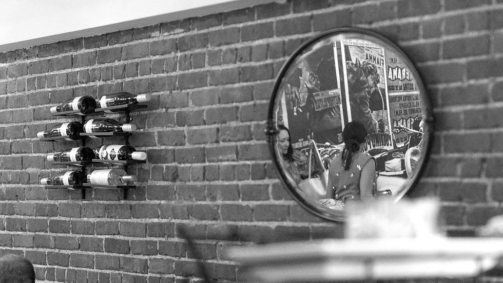 A mirror on a brick wall reflects a woman in a hat near vintage posters. Wine bottles are on a rack nearby. Black and white image.