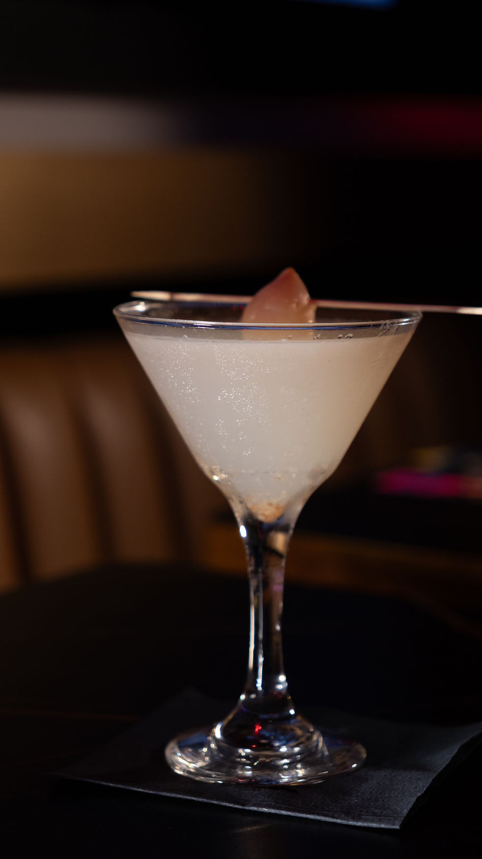 A clear cocktail in a martini glass garnished with a pink fruit, set on a dark table with a blurred background, conveying a classy mood.