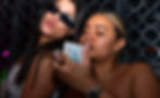 Two women in a dimly lit club. One wears sunglasses, puckering lips, the other holds a decorated phone, both showing amused expressions.