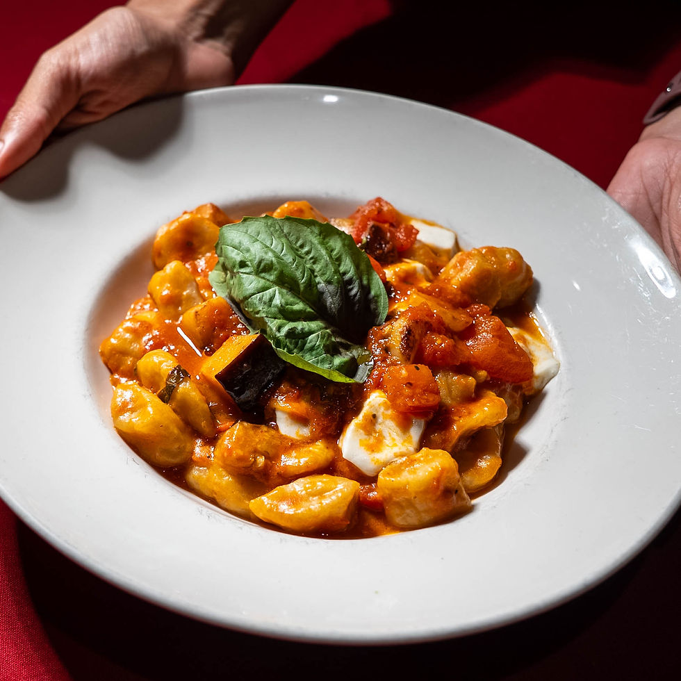 Plate of gnocchi with tomato and mozzarella, topped with basil. Hands holding the plate over a red tablecloth. Bright, appetizing colors.