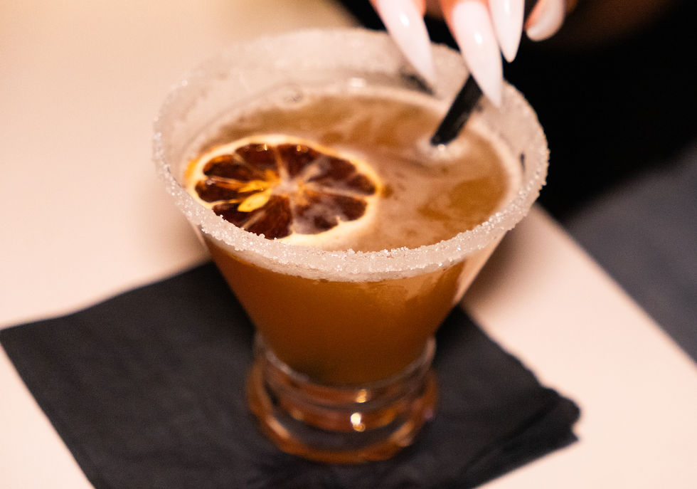 Cocktail in a sugar-rimmed glass with a dried orange slice on top. A hand with long nails stirs with a black straw on a dark napkin.