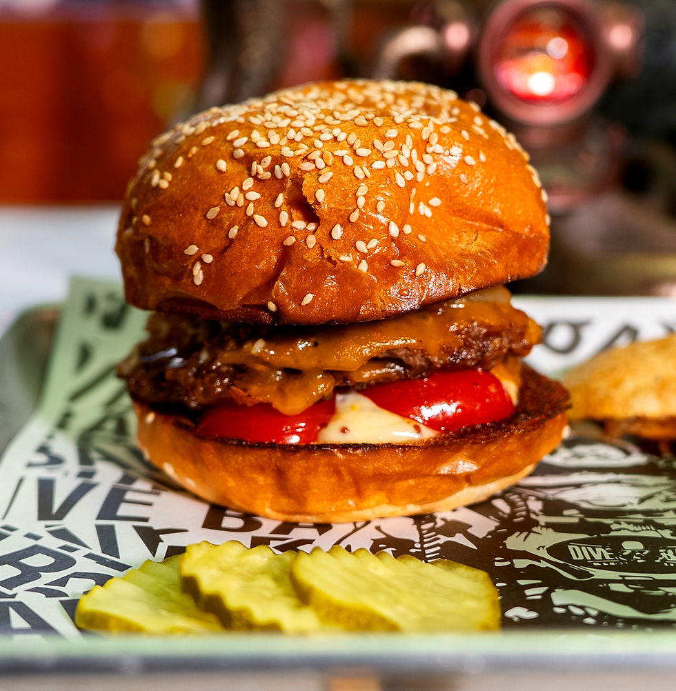 Close-up of a juicy cheeseburger with melted cheese, tomato, and sesame bun on a tray with pickles. Bold text in backdrop adds diner vibe.