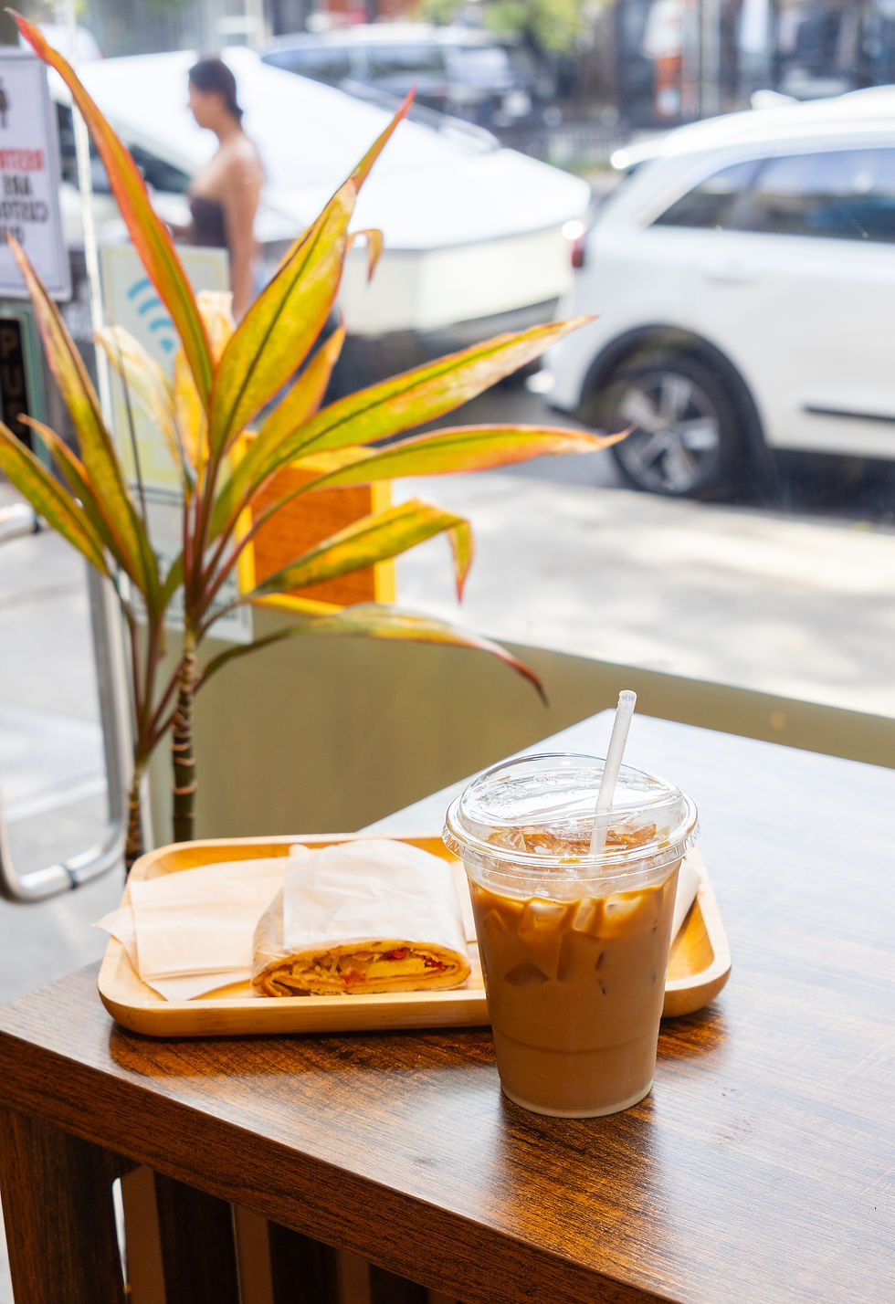 Iced coffee and wrapped sandwich on a wooden table, with a colorful plant indoors. A woman and cars are visible outside through the window.