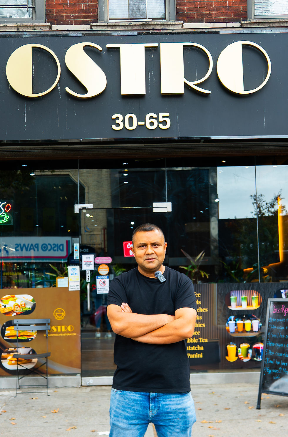 Man stands confidently with arms crossed outside a shop named OSTRO, address 30-65. Dark storefront with colorful signs and menu.