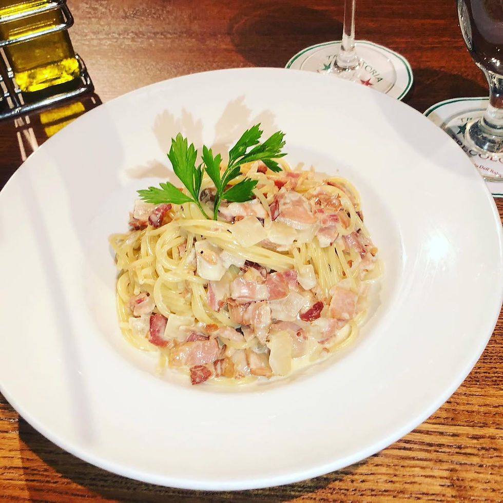 Pasta with creamy sauce, bacon bits, and parsley on a white plate. Set on a wooden table with oil, vinegar, and wine glasses nearby.