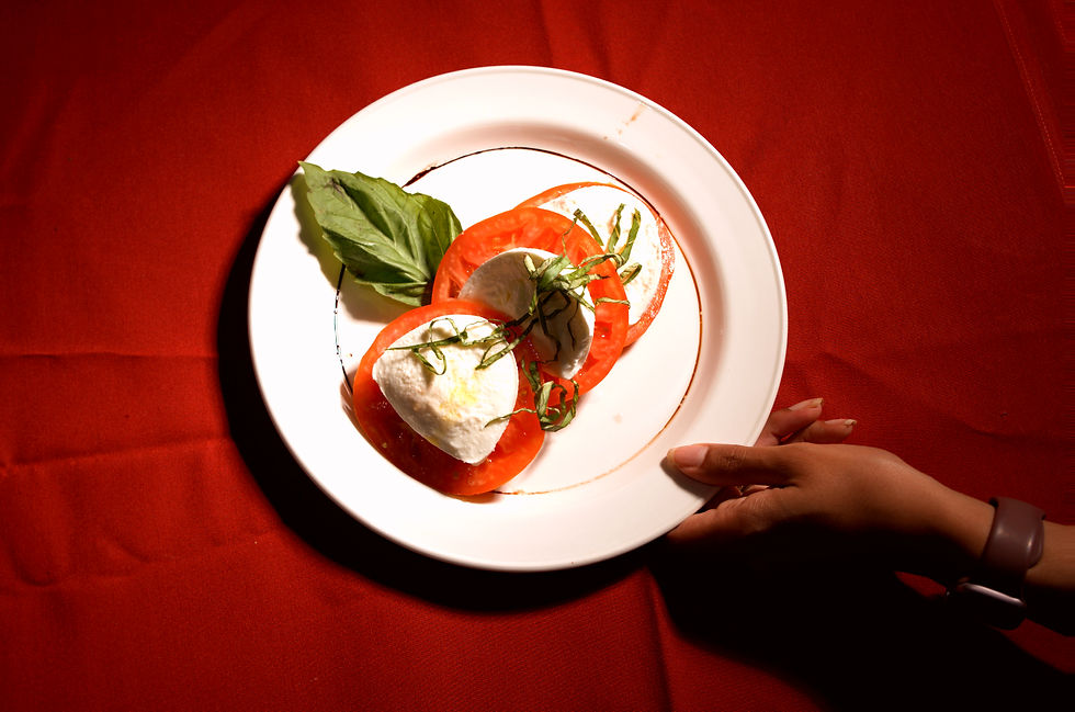 Caprese salad on a white plate with tomatoes, mozzarella, and basil. Held by a hand over a red tablecloth, creating a fresh and vibrant mood.