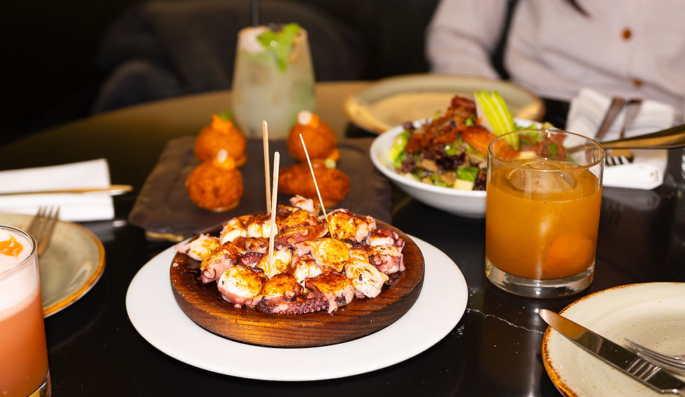 A table with octopus dish, fried appetizers, salad, and cocktails. Plates, utensils, and napkins are visible. Warm, inviting setting.
