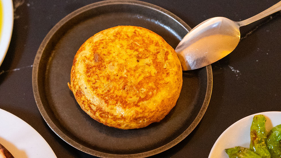 Round omelet on a dark plate with a spoon beside it. The omelet is golden-brown. White plates with food are partially visible.