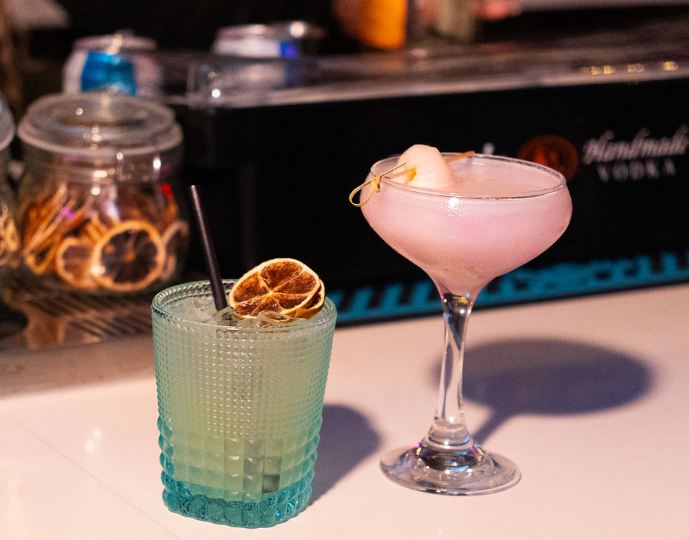Pink cocktail in a coupe glass with a peach garnish and green drink with dried citrus slice in a textured glass on a bar counter.