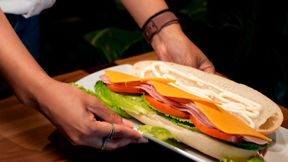 Hands holding a sandwich with lettuce, tomato, ham, cheese, and mayo on white bread, set against a dark leafy background.