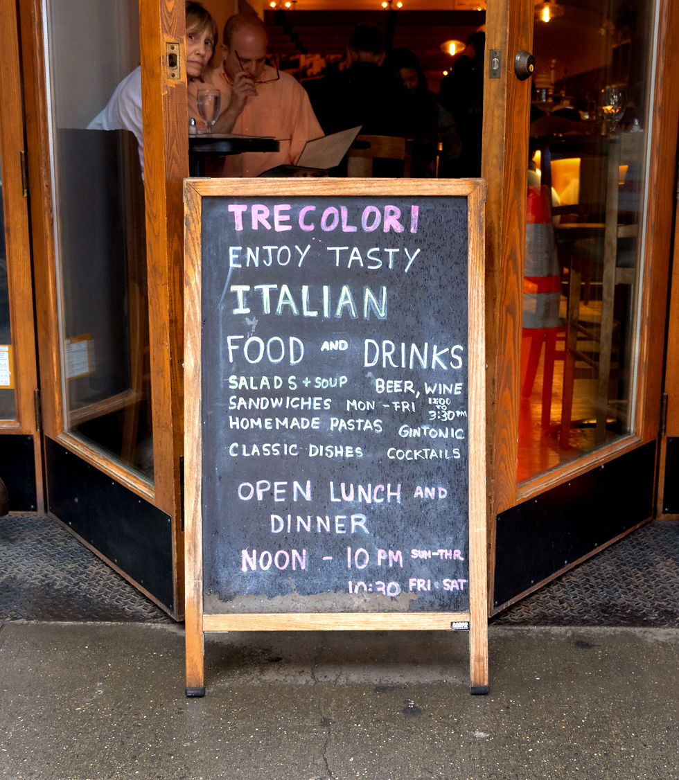 Chalkboard menu outside an Italian restaurant. It lists food, drinks, and hours. Two people are inside, one reading, in a cozy setting.