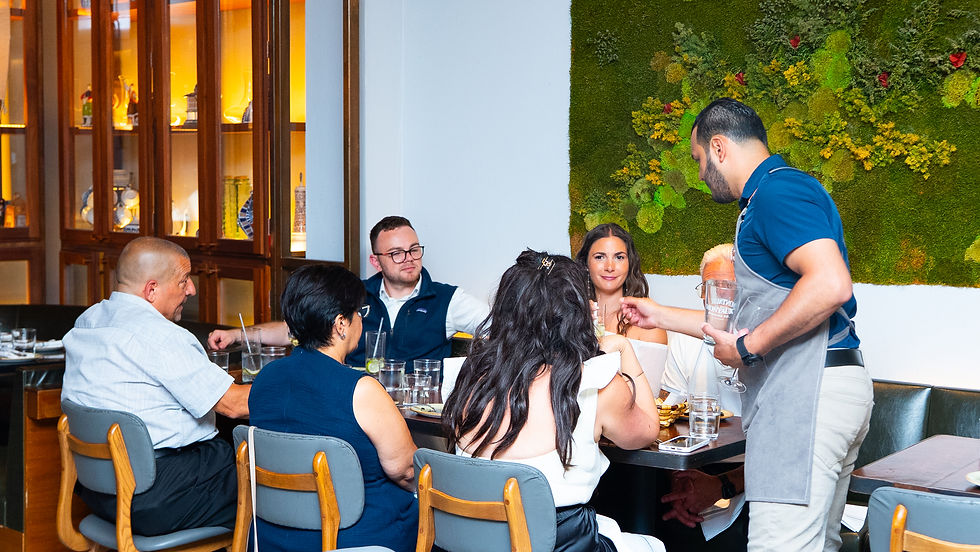 Server in blue shirt serves water to a group of six people dining at a restaurant. Wall with green moss art in the background.