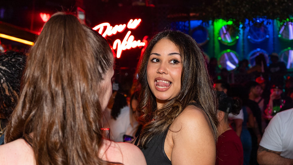 Women in a nightclub smiling with colorful lights and "Dare to Vibe" sign in background. Lively atmosphere, hair in ponytail, joyful mood.