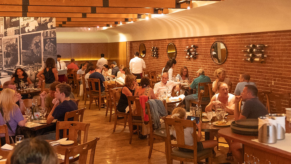 People dining in a cozy restaurant with brick walls and wooden décor. Mirrors and wine bottles adorn the walls. Warm, lively atmosphere.