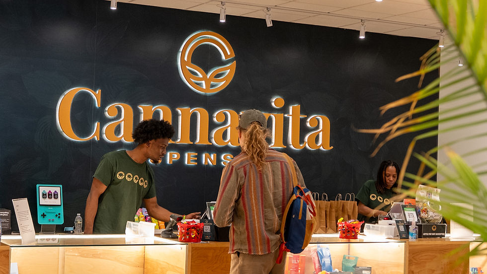 Two people behind a counter help a customer at a dispensary. The backdrop displays "Cannavita." Warm lighting and a casual setting.