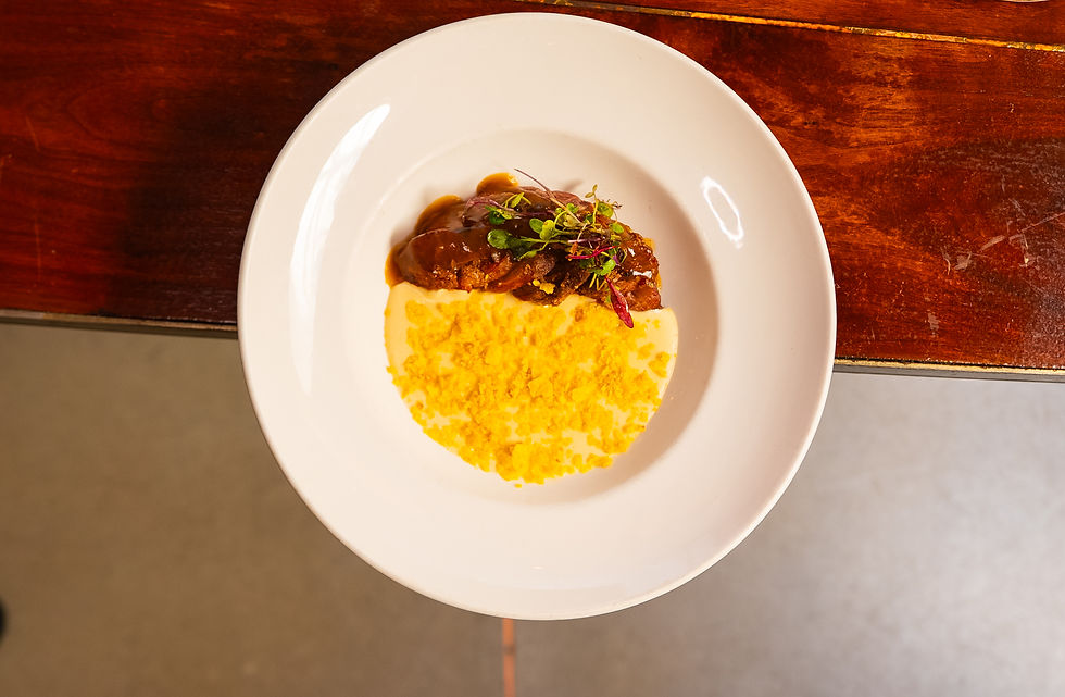 A white plate with sauce-covered meat, mashed potatoes, crumb topping, and microgreens, on a wooden table. Warm and appetizing presentation.