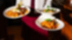 Server in white shirt and red tie holds three plates of pasta. Dishes feature vibrant sauces and garnishes. Warm indoor setting.