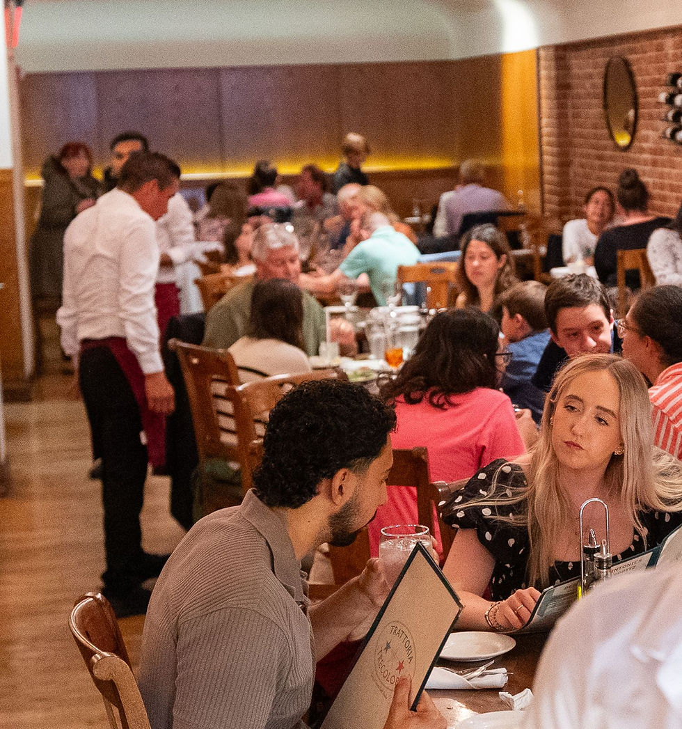Restaurant scene with people dining, a waiter serving. Patrons chat and review menus. Warm lighting, brick wall, casual and lively atmosphere.