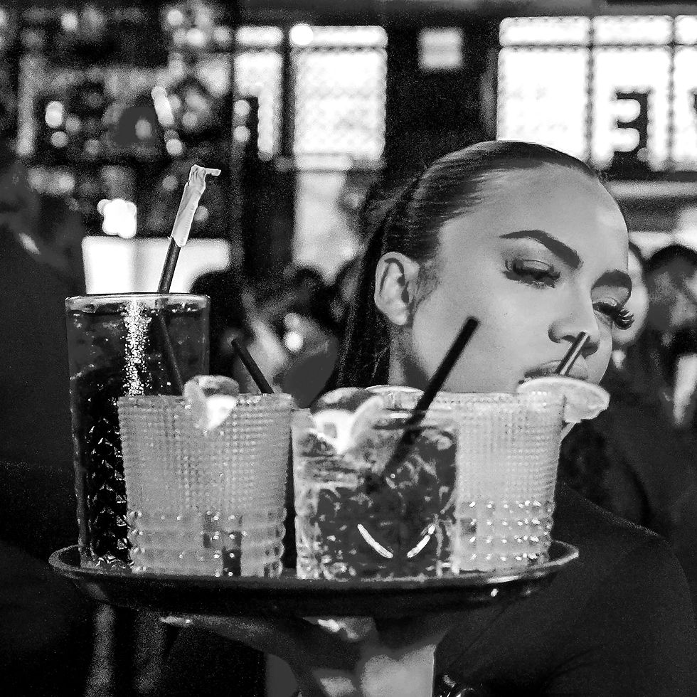 Server carrying a tray of drinks with limes and straws in a dimly lit bar. Background blurred with people and lights, creating a lively mood.