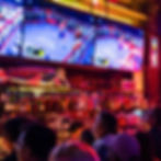 People watch a boxing match on bar TVs. Neon lights illuminate bottles and a mermaid statue. The scene is lively and vibrant.