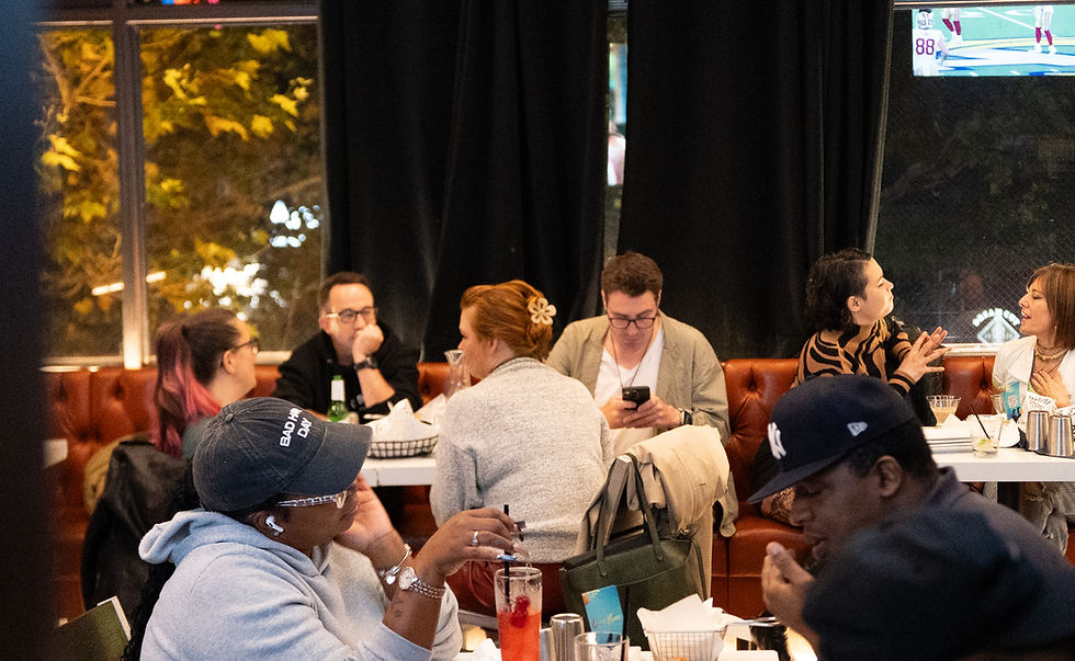 People sitting at a restaurant table; one uses a phone, others talk. Warm lighting and drinks on the table create a social, relaxed mood.