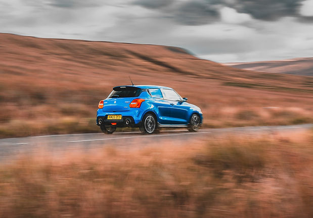 Suzuki Swift Sport Review - rear three quarter panning shot on a moorland road with the Peak District mountain range as the backdrop