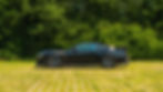 Side profile of the Clive Sutton CS800DH Mustang in black, showcasing its carbon fibre bonnet vents, side skirts, large rear wing, and Vossen wheels, parked on grass with dense green foliage in the background.